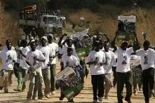 A group of young people conversing on the street for their party of choice, wearing party regalia and holding posters. A Truck packed with supporters holding a Zimbabwean flag follows them in the background