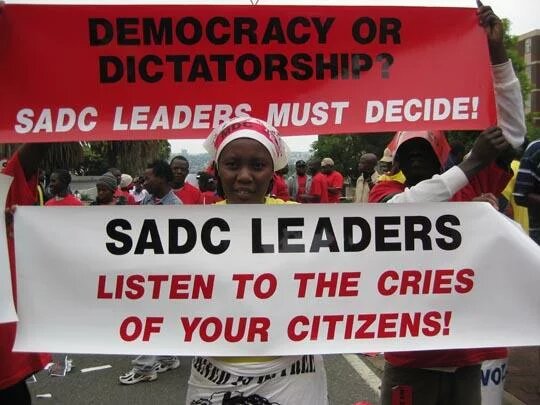 A protester holds up a poster that reads “SADC Leaders Listen to the cries of your citizens”. Above her, is a poster that reads “Democracy or Dictatorship? SADC leaders must decide!”