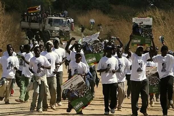 A group of young people conversing on the street for their party of choice, wearing party regalia and holding posters. A Truck packed with supporters holding a Zimbabwean flag follows them in the background